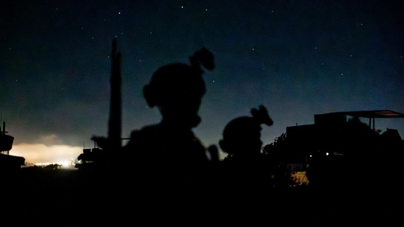 Silhouettes of soldiers wearing helmets with mounted equipment against a night sky with stars, with distant lighting illuminating the horizon at what appears to be a military outpost or forward operating base.