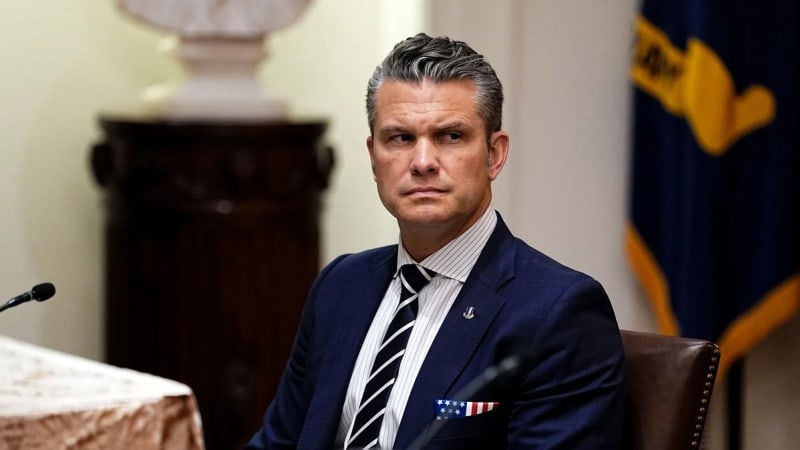 A man in a navy blue suit with a striped tie and American flag pocket square sits at a formal dining table in what appears to be a government building, with a microphone and official flag visible in the background.