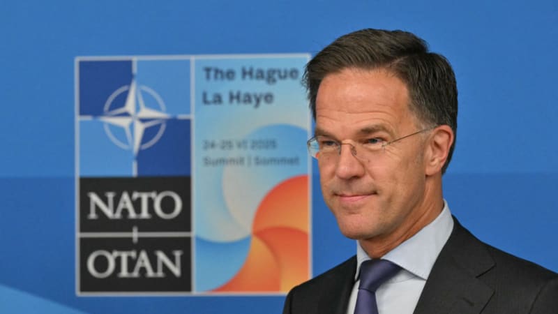 A man in a dark suit and glasses stands in front of a NATO summit backdrop for The Hague 2025, featuring the NATO/OTAN logo on a blue background.