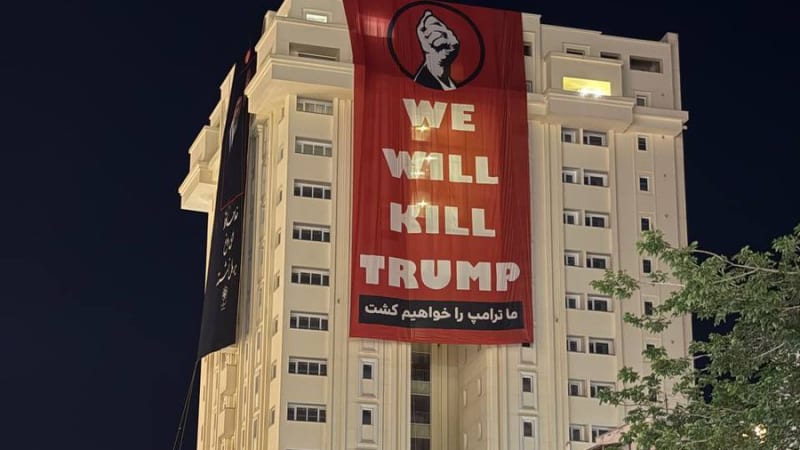 A large red banner hung on a building at night reads 'WE WILL KILL TRUMP' in English with Persian text below, alongside a raised fist graphic. Black banners with Persian/Arabic script are also visible on the building, likely in Iran.