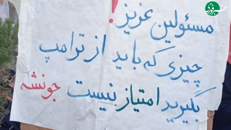 A handwritten protest sign in Persian (Farsi) held up at a demonstration. The text addresses officials and references Trump, stating that what should be taken from Trump is not concessions but his life/spirit. People and a cleric figure are visible in the background.