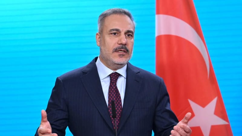 A man in a dark suit and patterned tie gestures while speaking at a podium, with a Turkish flag visible in the background against a blue backdrop.
