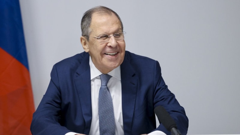 A smiling elderly man in a navy blue suit and patterned tie sits at a desk holding papers, with a microphone in front of him and a Russian flag visible in the background.