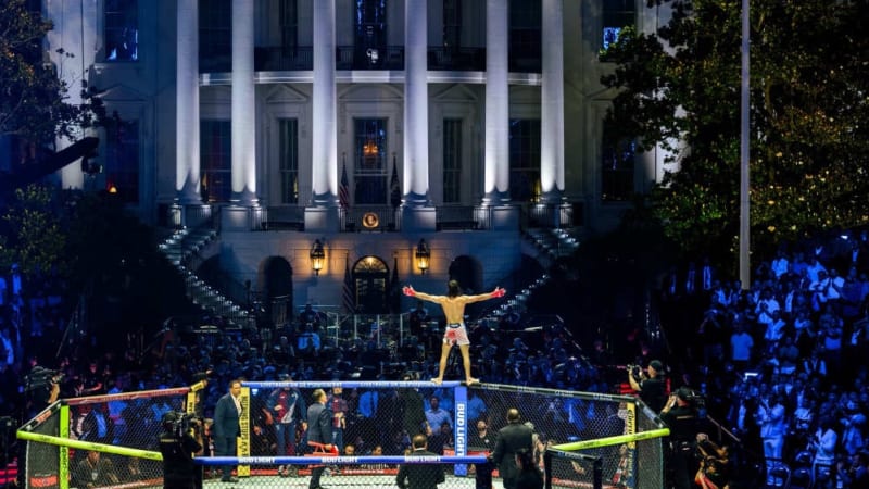 A shirtless fighter stands on top of a UFC octagon cage with arms outstretched, facing the illuminated White House South Portico at night, with a large crowd gathered for what appears to be a UFC event held on the White House South Lawn.