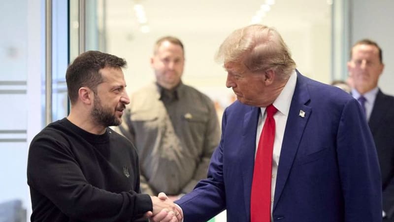 Two men shake hands in a formal indoor setting, with security personnel visible in the background. One wears a black sweater and olive pants, while the other wears a blue suit with a red tie.