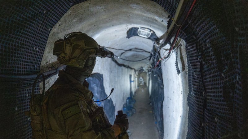 An armed soldier in full tactical gear including helmet with night vision equipment stands inside an underground tunnel with concrete walls, wiring, and water bottles visible in the corridor, consistent with imagery from Israeli military operations in Gaza tunnels.