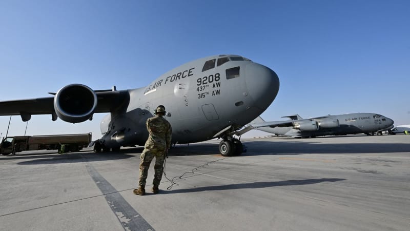 A U.S. Air Force crew member in camouflage uniform stands on the tarmac in front of a C-17 Globemaster III (tail number 9208, 437th AW / 315th AW), with a second C-17 visible in the background.