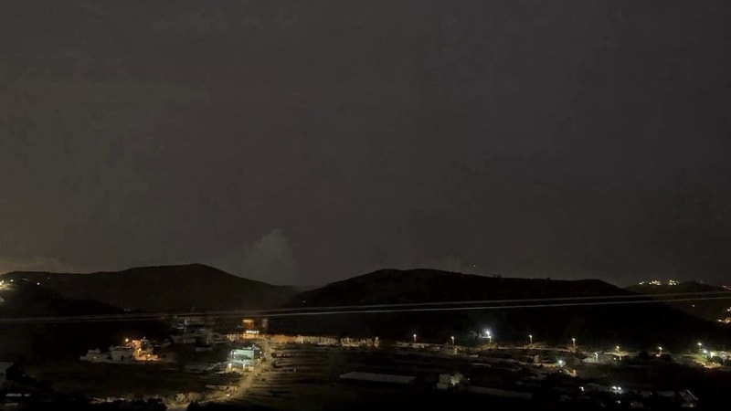 Nighttime photograph of a small coastal or valley town with scattered lights, silhouetted hills in the background, and a dark sky with faint wispy clouds.