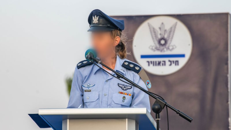 A female Israeli Air Force officer with blurred face speaks at a podium during an official ceremony, with the Israeli Air Force (חיל האוויר) emblem visible on a banner behind her.