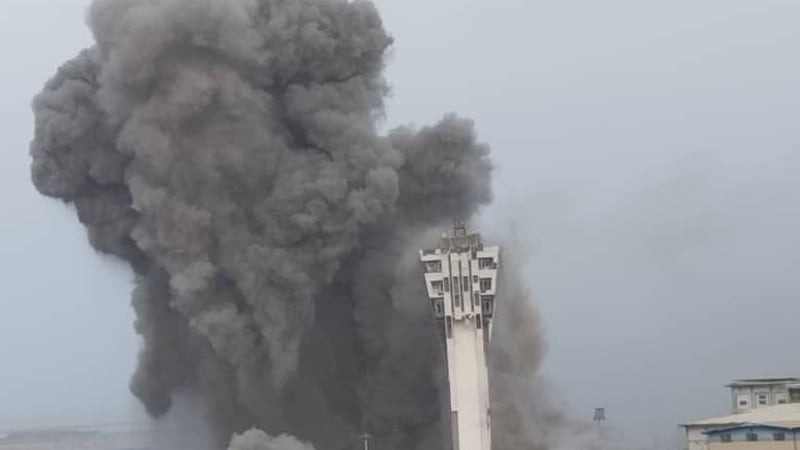 A large explosion sends a massive plume of smoke and debris into the sky near a tall tower structure, likely an airport control tower, in what appears to be an airstrike in a conflict zone, possibly Yemen.