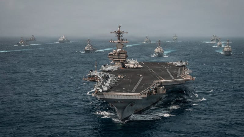 A U.S. Navy aircraft carrier with fighter jets on its flight deck leads a large naval formation of warships sailing in open ocean, likely during a military exercise or show of force.