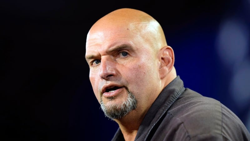 Close-up portrait of a bald man with a goatee speaking, wearing a dark shirt against a dark blue background.