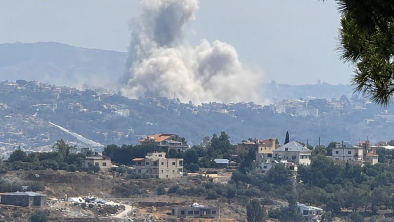 A large plume of smoke rises from an airstrike or explosion over a hillside town, likely in southern Lebanon, with residential buildings and vegetation in the foreground.