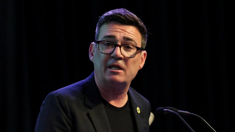 A man wearing glasses, a dark blazer, and a black t-shirt with a small lapel pin speaks at a microphone against a dark background.