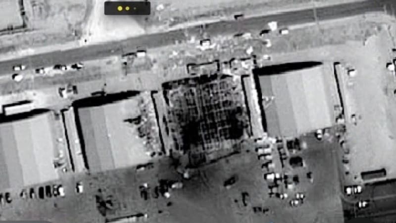 Satellite imagery showing damaged or destroyed warehouse/hangar structures at what appears to be a military or industrial facility, with one building visibly collapsed or heavily damaged. A watermark logo is visible in the upper left corner.