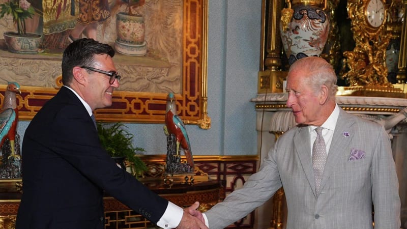 Two men in formal suits shaking hands in an ornate palace room decorated with paintings, gilded furniture, and decorative porcelain. The setting appears to be a royal or state reception room.
