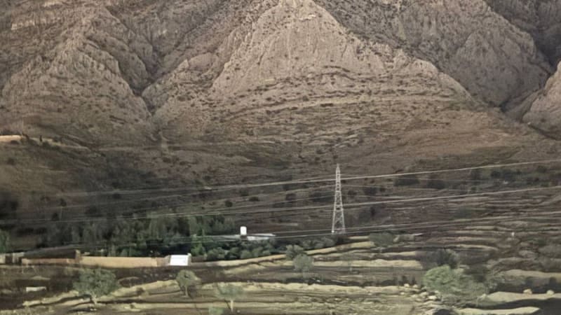 A plume of smoke rises from behind a rugged mountain range at dusk or night, with a power transmission tower visible in the foreground valley, suggesting an airstrike or explosion in a mountainous region.