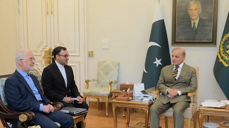 A diplomatic meeting in an official Pakistani government setting, showing three men seated in formal attire with a Pakistani flag and a portrait of Muhammad Ali Jinnah in the background.