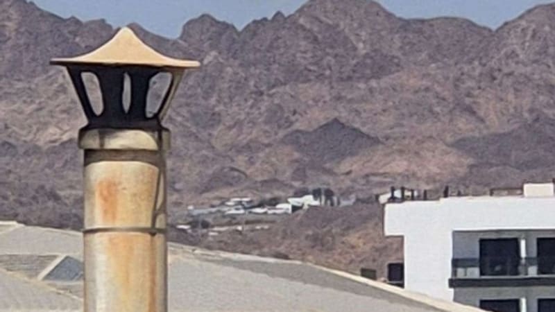 A plume of smoke rising above arid mountains, photographed from an urban area with buildings and a decorative chimney or lamp post in the foreground, likely depicting an explosion or airstrike in a Middle Eastern setting.