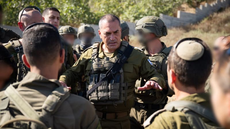 A senior Israeli Defense Forces (IDF) officer wearing tactical gear and binoculars briefs a group of soldiers in olive-green uniforms, some wearing helmets and one wearing a kippah, in an outdoor field setting.