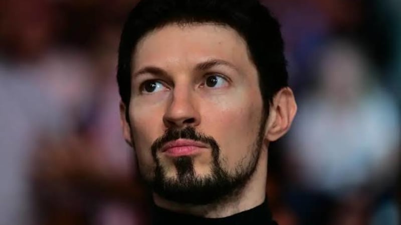 Close-up portrait of a man with dark hair and a beard wearing a black turtleneck, looking upward with a serious expression, with a blurred audience in the background.