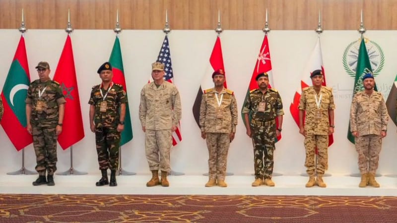 Military officials from multiple nations stand in a formal group photo on a stage, each positioned in front of their respective national flags, including those of Pakistan, Morocco, the United States, Bahrain, Oman, Qatar, Saudi Arabia, Sudan, Jordan, the United Kingdom, and Australia, likely at a multinational military conference or exercise.