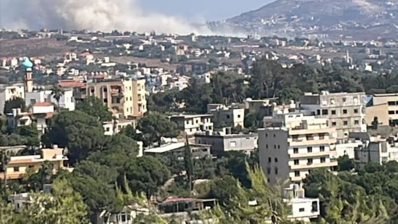 A plume of smoke rises in the distance over a hilly Lebanese town with residential buildings, trees, and a mosque minaret visible in the foreground, suggesting an airstrike or explosion.
