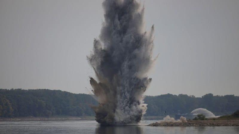 A large explosion sends a column of water, debris, and smoke high into the air from a river or lake, with forested hills visible in the background, likely a controlled detonation of unexploded ordnance.