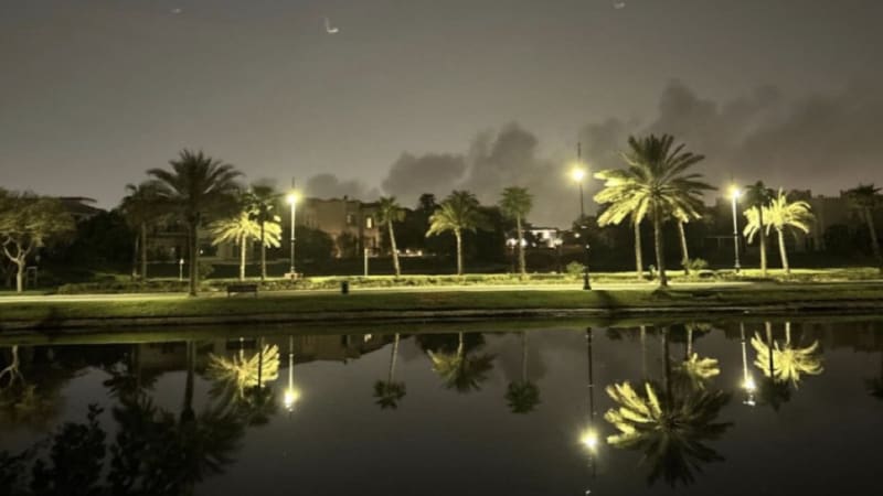 Nighttime photo of a palm tree-lined waterfront promenade with street lights reflecting in calm water, with dark smoke plumes visible rising in the background sky, suggesting nearby fires or explosions in what appears to be a Middle Eastern city.