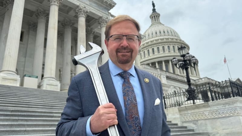 A man in a blue suit holding a large adjustable wrench poses on the steps of the U.S. Capitol building in Washington, D.C.
