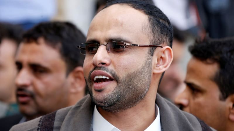 Close-up photo of a man wearing glasses and a white shirt with a brown shawl, speaking while surrounded by other men in what appears to be a public gathering.