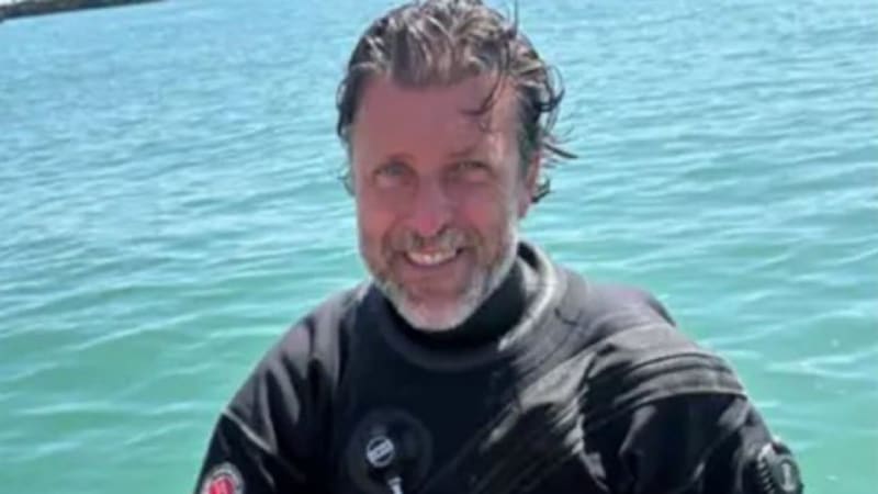 A smiling man in a black diving wetsuit sits on an inflatable boat, holding what appears to be an old bottle recovered from underwater. The turquoise sea is visible in the background.