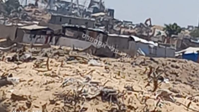 A devastated urban landscape showing destroyed buildings and rubble in the background with makeshift shelters and tents in the middle ground, taken from behind a sandy embankment. Hebrew text watermark visible. Photo taken with a Samsung Galaxy A32, likely depicting destruction in Gaza.
