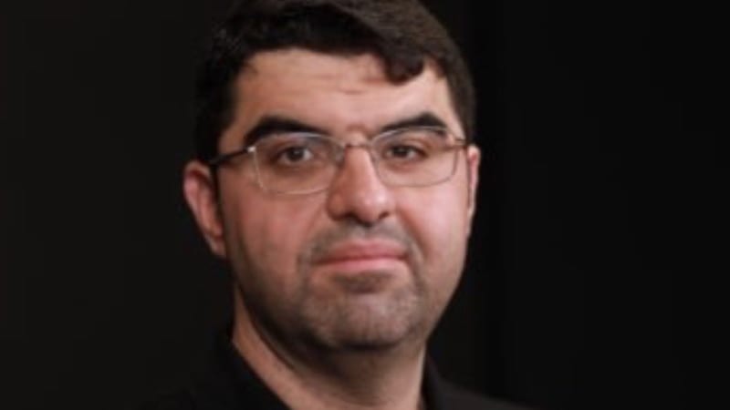 Portrait photograph of a middle-aged man with short dark hair, glasses, and a goatee, wearing a black polo shirt against a dark background.