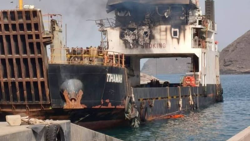 A damaged and burning vessel named 'TIHAMAH' docked at a port, with black smoke rising from its superstructure showing significant fire and blast damage, consistent with a conflict-related attack.