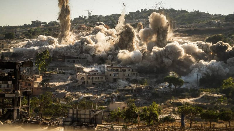 Multiple large explosions send plumes of dust and debris into the air across a heavily damaged urban area, with destroyed buildings and rubble visible in the foreground, consistent with an airstrike or bombardment in a conflict zone.