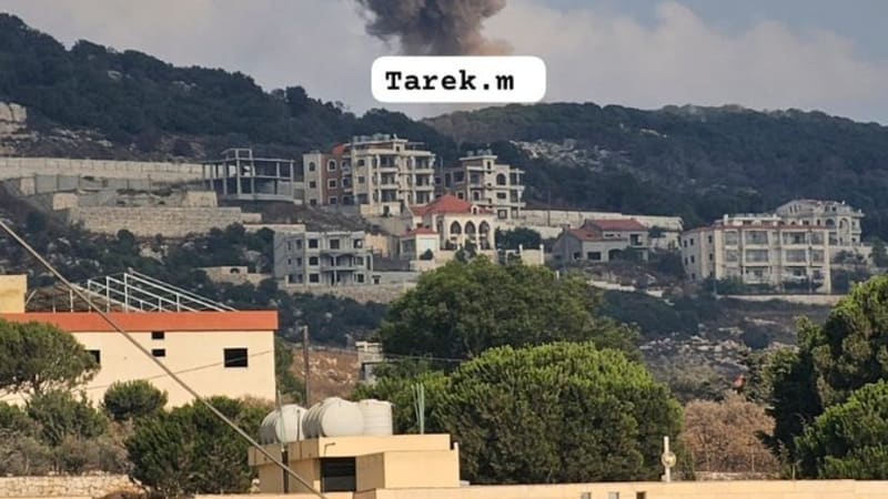 A large column of smoke rises from an airstrike behind a hillside town, likely in southern Lebanon, with residential buildings visible in the foreground and on the hillside. The image has a watermark reading 'Tarek.m'.