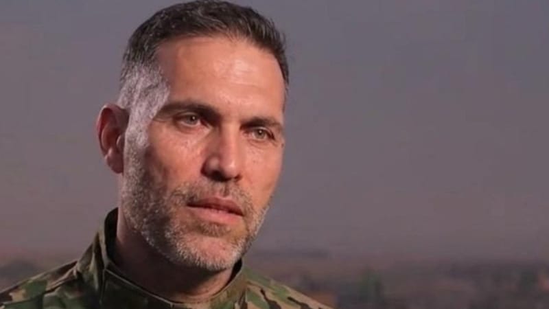 Close-up portrait of a middle-aged man wearing military camouflage uniform, with short graying hair and stubble, photographed outdoors with a hazy background.
