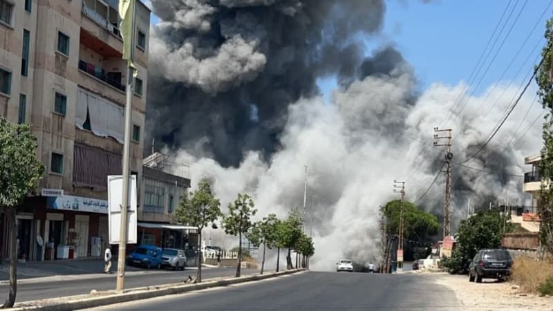 A massive plume of dark smoke rises from an explosion in a Middle Eastern urban area, with residential buildings, parked cars, and Arabic signage visible along a street.