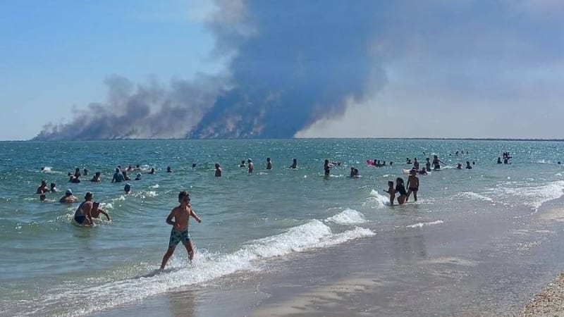 Beachgoers swim and wade in the sea while a massive wildfire smoke plume rises from the distant shoreline, creating a stark contrast between leisure and disaster.