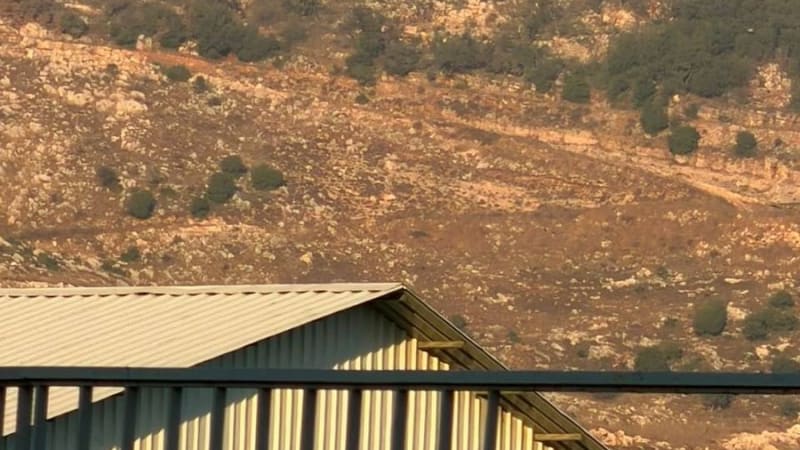 A large plume of brown and gray smoke rises from a hillside covered with sparse vegetation and rocky terrain, viewed from behind a corrugated metal roof structure, consistent with an airstrike or explosion in a Middle Eastern landscape.