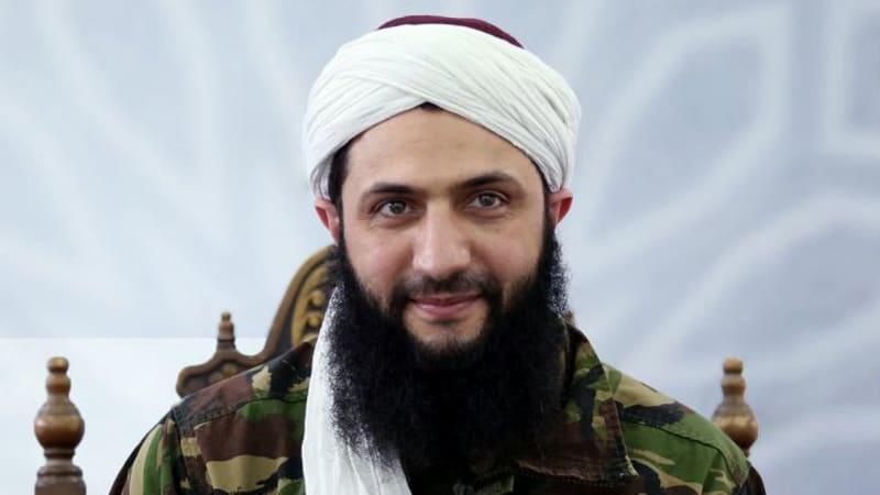 A bearded man wearing a white turban and military camouflage jacket, seated and looking at the camera with a slight smile.