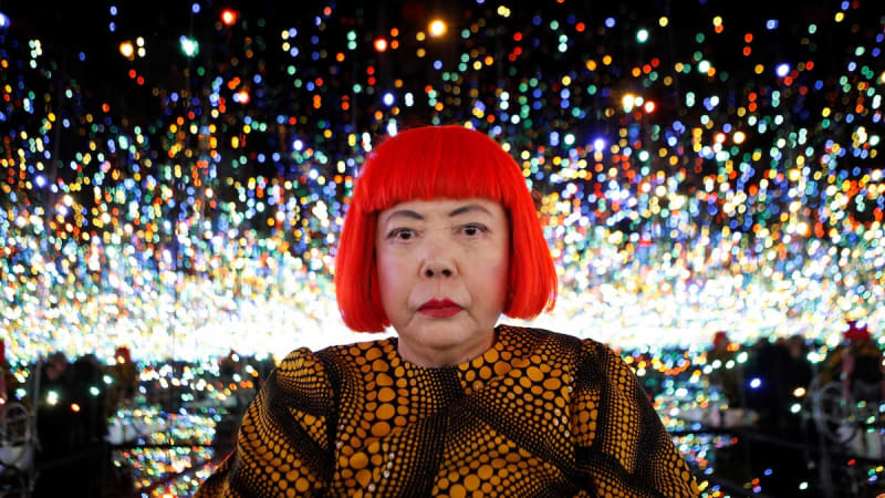 An elderly woman with a bright red bob wig and yellow-and-black polka dot clothing poses inside an immersive infinity mirror room filled with countless multicolored lights, characteristic of Yayoi Kusama's iconic art installations.