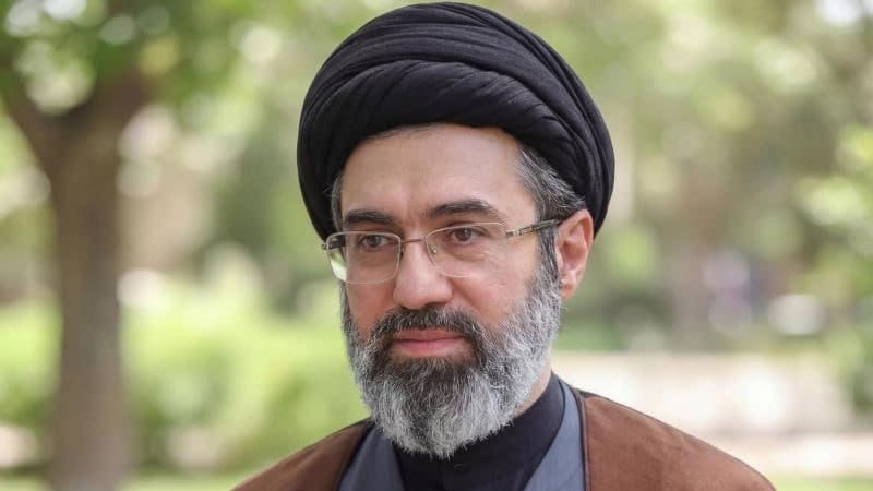 Portrait of a middle-aged man wearing a black turban, glasses, and clerical robes, with a gray beard, photographed outdoors with a blurred green background.