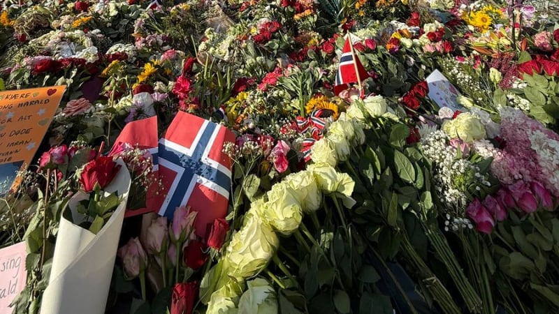 A massive floral memorial with Norwegian flags and handwritten notes covers the grounds in front of the Royal Palace in Oslo, Norway, with the royal standard flying at the palace.