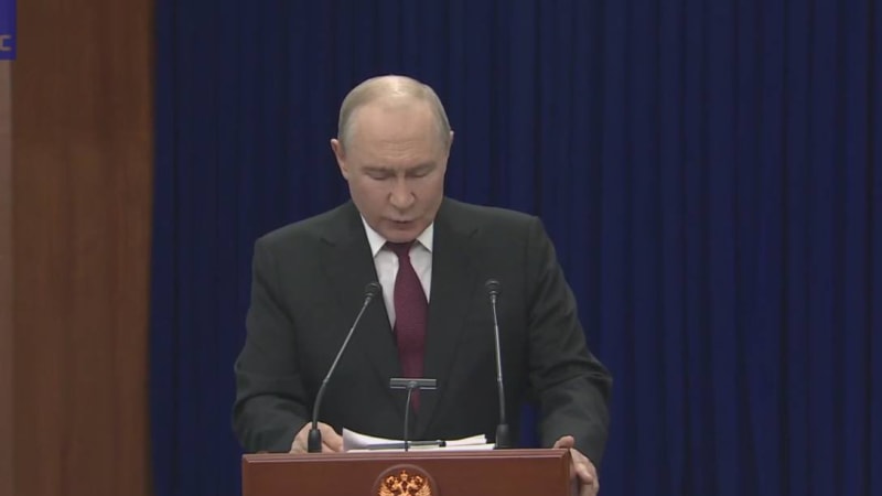 A man in a dark suit and red tie speaks at a podium bearing the Russian state emblem, with a blue curtain backdrop.