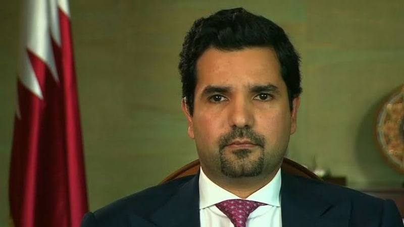 A man in a dark suit and patterned tie sits in front of a Qatari flag, appearing to be in an official or diplomatic setting during a television interview.