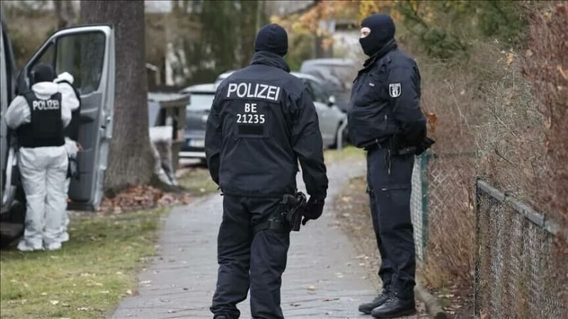 German Berlin police officers in dark uniforms and balaclavas during a raid or operation, with a forensic investigator in white coveralls visible in the background near a van.