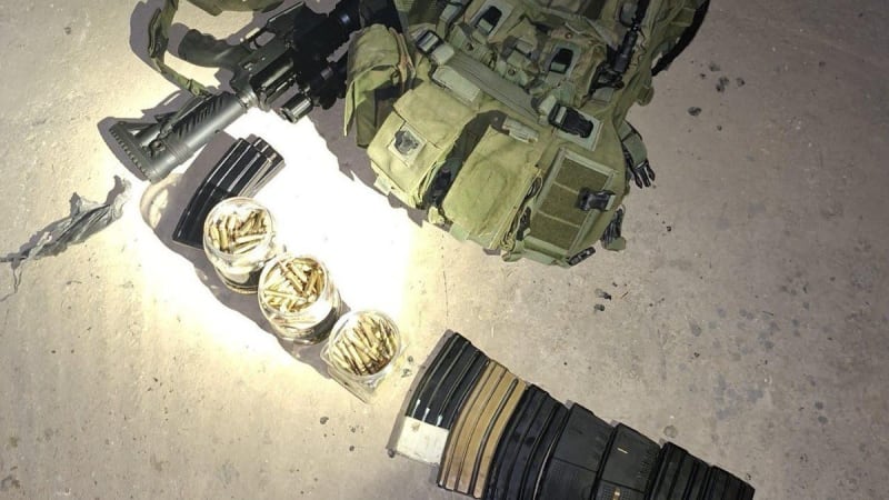 Seized or displayed military-style equipment on a concrete floor, including an M4/AR-15 type rifle with optic, a green tactical plate carrier vest, multiple rifle magazines, and containers of loose ammunition.