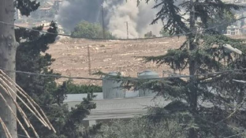 A large plume of smoke rises from an explosion in an urban area, viewed through trees and power lines from a nearby hillside or neighborhood.